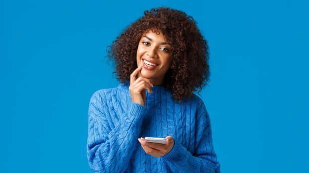 Frau hält ihr Smartphone und lächelt in die Kamera. 
